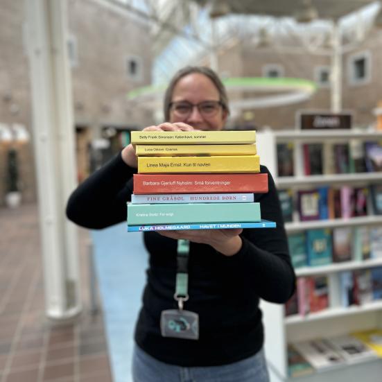 Tine står blandt fyldte bogreoler på Hovedbiblioteket. Hun holder syv af de omtalte bøger i denne artikel frem for sig i en stak med bogryggene frem. Der er fokus på bogryggene, mens både Tine og resten af baggrunden, som viser biblioteksrummet, fremstår let sløret. Man kan ane, at Tine smiler.