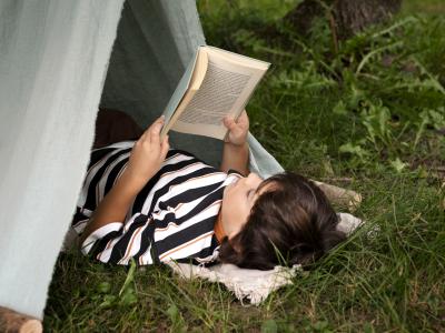Barn, der ligger halvt inde i et telt udenfor og læser.
