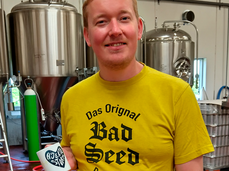 Frederik fra Bad Seed Brewing står foran ølkedler med en "Hjertestorm-kop" i hånden.