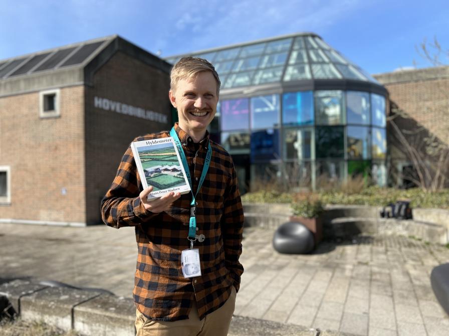 Mathias står i bibliotekshaven ved Hovedbiblioteket. Han holder bogen "Hyldemeter" i den ene hånd og har den anden hånd i lommen. Bag Mathias kan man se glashjørnets farverige vinduer. Mathias smiler og kigger lidt til højre for kameraet. Himlen er blå og solen skinner.